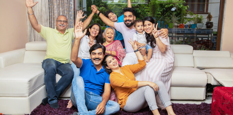 Happy multi-generational Indian family smiling and waving together in a living room, representing successful family visa immigration and reunion.